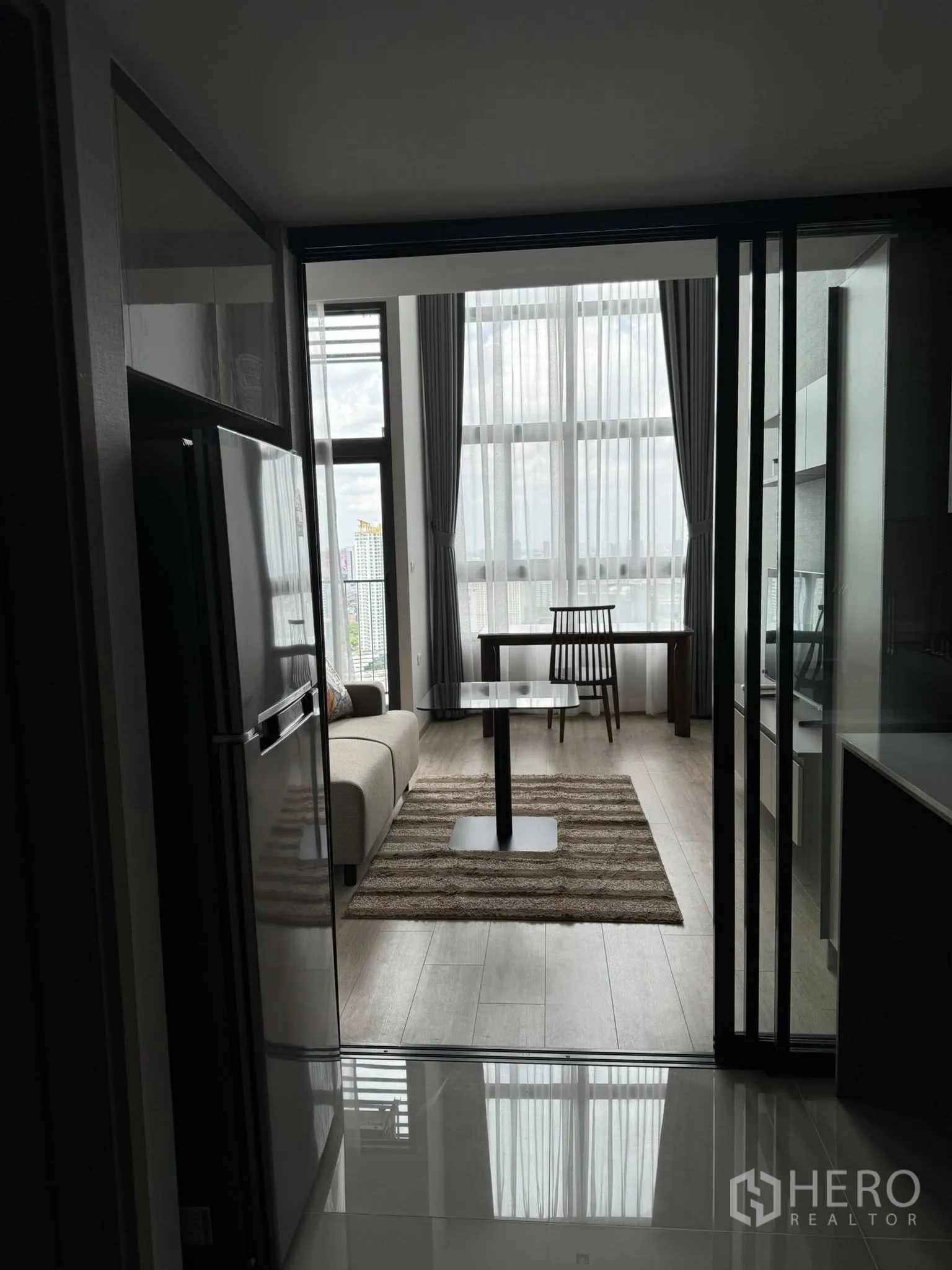 Condominium for rent in Huai Khwang, Bangkok - Modern condo living area with floor-to-ceiling windows, sofa, rug and dining table viewed from the kitchen.