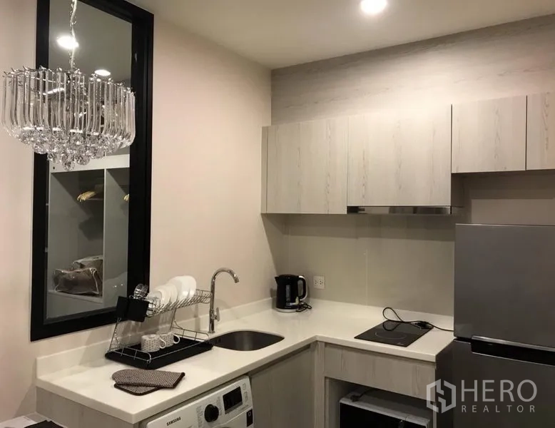 Condominium for rent in Huai Khwang, Bangkok - L-shaped kitchen with light wood cabinets, sink, cooktop, fridge, washing machine and a crystal chandelier.
