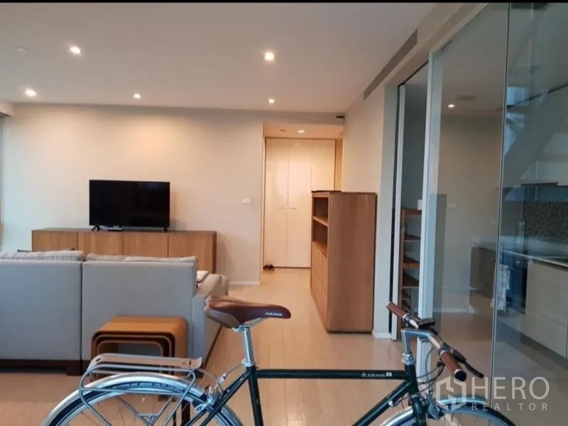 Condominium for rent or sale in Watthana, Bangkok - Spacious lounge with sectional sofa and TV, with sliding glass doors opening to a fitted kitchen.