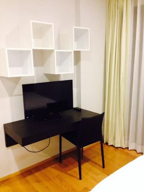 公寓 出租在Phaya Thai, Bangkok - 工作角落，壁挂书桌、平板电视、座椅与方格置物架，旁有落地窗帘。