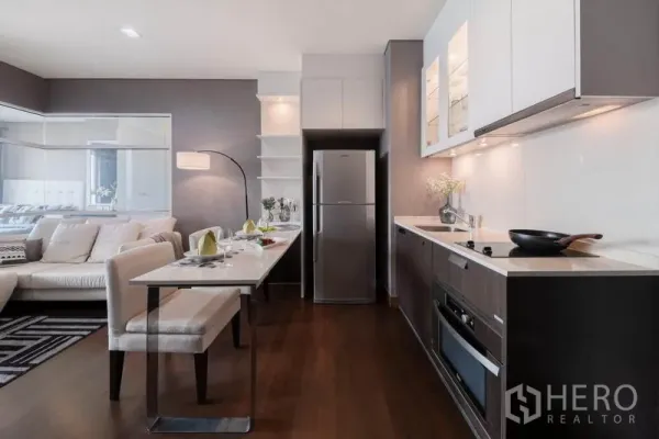 公寓 出租在Watthana, Bangkok - 现代直线型厨房，内置家电与烤箱，旁设早餐桌。