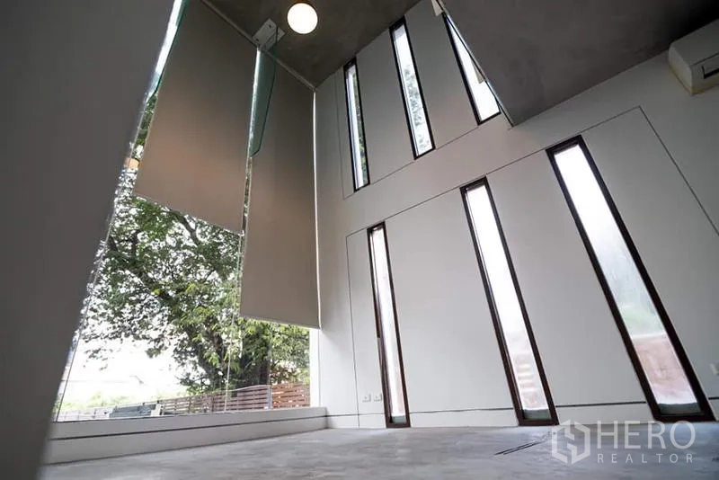 ทาวน์เฮาส์ ให้เช่าหรือขายในPhra Khanong, Bangkok - โถงเพดานสูงพร้อมหน้าต่างทรงยาวหลายบานและม่านม้วน มองเห็นต้นไม้ด้านนอก