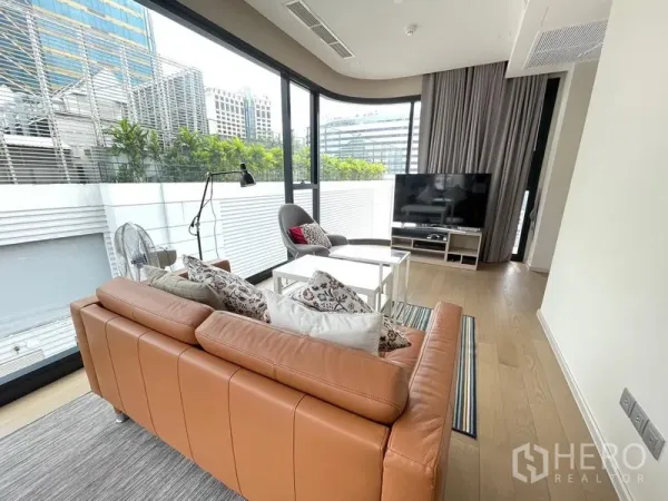 Condominium for rent or sale in Watthana, Bangkok - Bright living room with tan leather sofa, TV stand and floor‑to‑ceiling corner windows overlooking greenery and the city.