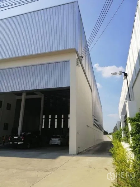 Warehouse for rent in Bang Phli, Samut Prakan - Exterior side view with tall metal cladding, wide driveway and a security camera.