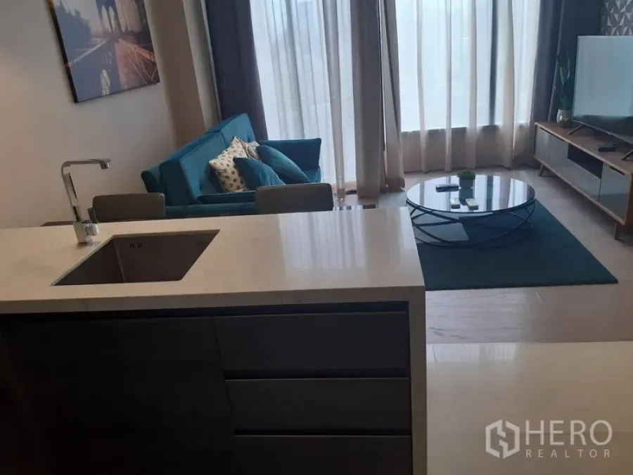 Condominium for rent or sale in Watthana, Bangkok - Open-plan living area with teal sofa, round coffee table, TV console and kitchen island beneath floor-to-ceiling windows.