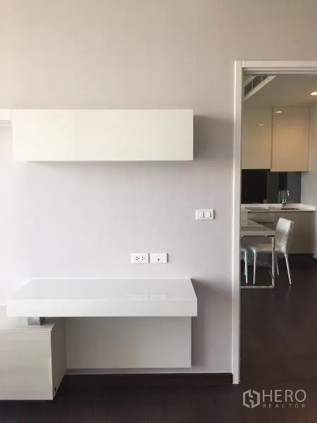 公寓 出租在Watthana, Bangkok - 极简墙面搭配悬空置物架，可见厨房与用餐区一角。