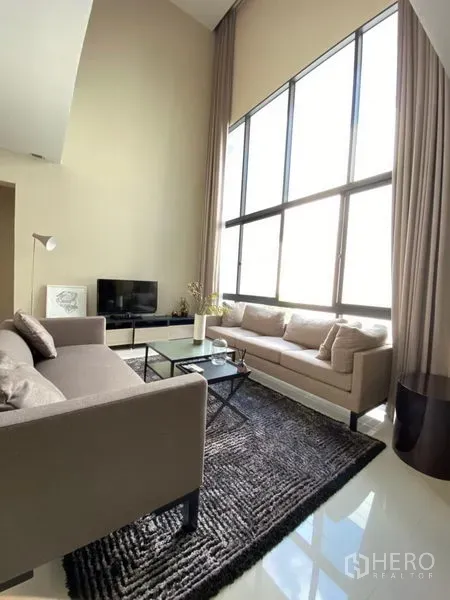 Detached House for rent in Huai Khwang, Bangkok - Living room with double-height ceiling, floor-to-ceiling windows, neutral sofas, rug, coffee table and TV.