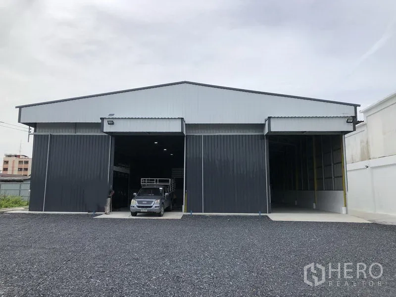 货栈 出租在Pathum Thani - 金属夹芯板外墙的仓库外观，双高卷帘门与碎石装卸场地