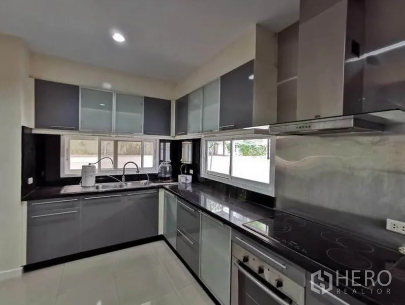 Detached House for rent in Bang Kapi, Bangkok - Modern U‑shaped kitchen featuring glossy cabinets, black countertops, oven, cooktop, hood and window lighting.