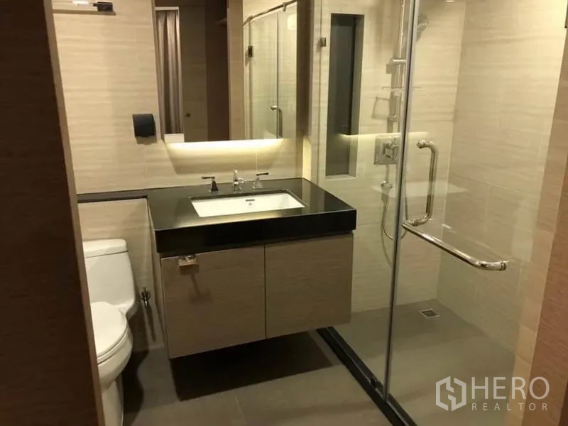 Condominium for rent in Pathum Wan, Bangkok - Modern bathroom with glass-enclosed shower, vanity with black countertop and toilet.