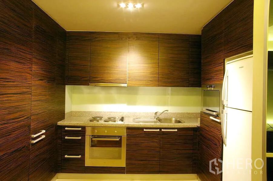 公寓 出租在Watthana, Bangkok - L形厨房，木纹橱柜、石材台面，嵌入式烤箱、灶台、水槽与冰箱。