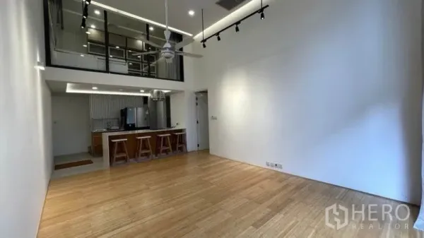 Condominium for rent in Watthana, Bangkok - Double-height living room with mezzanine glass wall, track lighting, ceiling fan, and open kitchen island with stools on warm wood flooring.