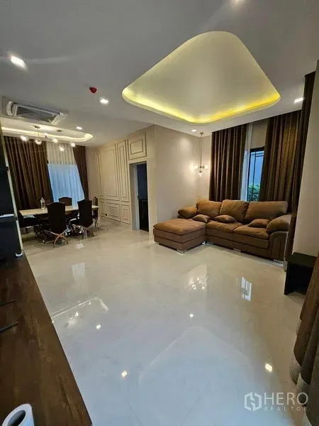 Detached House for sale in Mueang Pathum Thani, Pathum Thani - Spacious living room with L-shaped sofa, cove ceiling lights and large curtained windows.