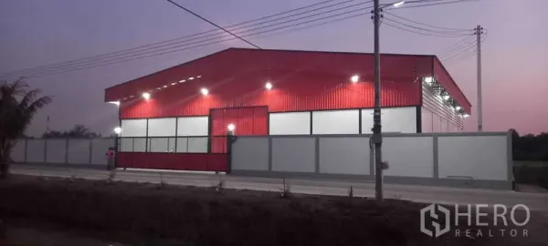 货栈 出租或出售在Khlong Luang, Pathum Thani - 黄昏街景，红色仓库立面与围墙，外部照明明亮。