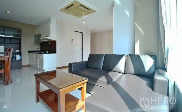 Condominium for sale in Watthana, Bangkok - Sunlit lounge with L-shaped leather sofa, coffee table and open kitchen beneath ceiling air conditioning.