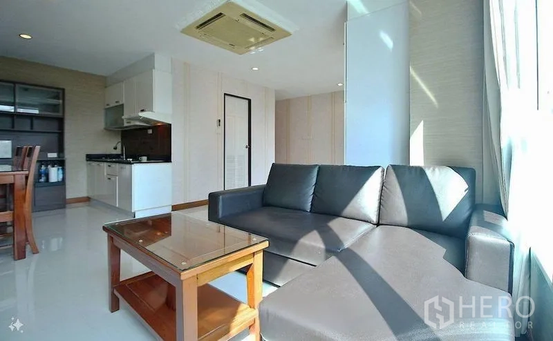 Condominium for sale in Watthana, Bangkok - Sunlit lounge with L-shaped leather sofa, coffee table and open kitchen beneath ceiling air conditioning.