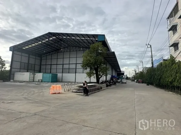 Warehouse for rent in Samut Prakan - Side view of the warehouse with covered loading bay, tree, and access road with power lines.