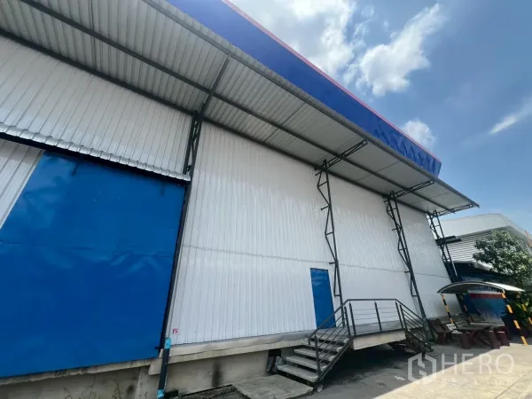 Warehouse for rent in Lat Krabang, Bangkok - Covered loading bay with a large blue sliding door, metal awning and dock-height stairs along the warehouse.