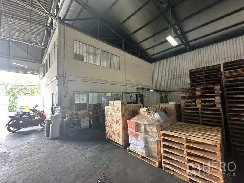 Warehouse for rent in Suan Luang, Bangkok - Corner storage area with pallets, boxes and a mezzanine office under a metal roof.