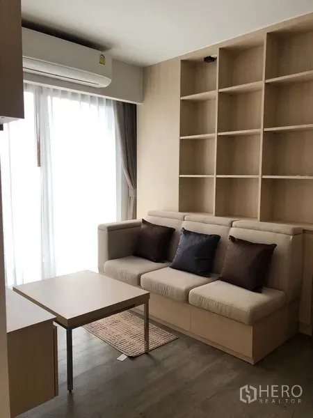 Condominium for rent or sale in Yan Nawa, Bangkok - Bright living area with beige sofa, built-in shelving, and floor-to-ceiling window with sheer curtains.