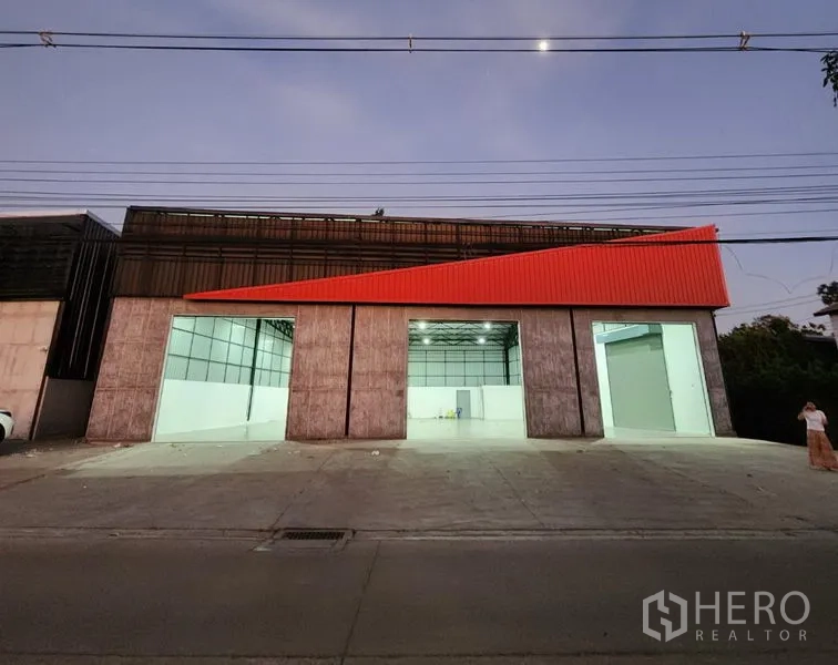 Warehouse for rent in Pak Kret, Nonthaburi - Dusk exterior with open bays revealing a bright warehouse interior beneath a high steel roof.