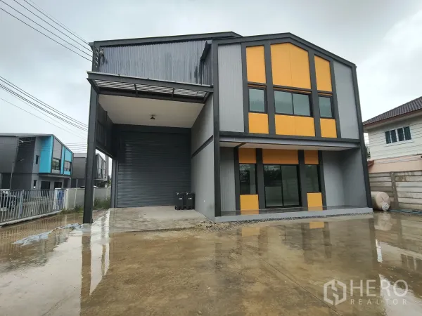 货栈 出租或出售在Bang Yai, Nonthaburi - 现代灰橙配色仓库外观，带两层办公区、遮雨装卸区、卷闸门与宽阔混凝土前院。