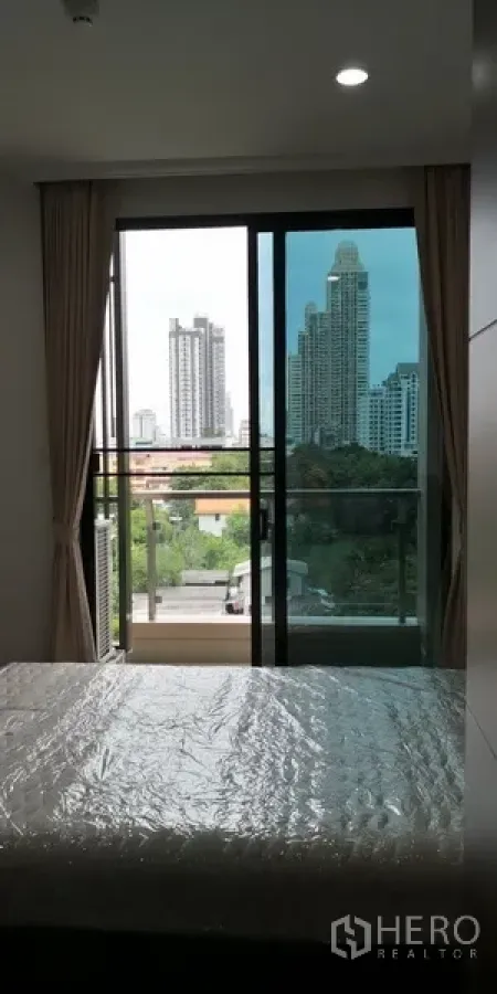 Condominium for rent in Sathon, Bangkok - Bedroom opening to a glass balcony with skyline and treetop views, framed by floor‑to‑ceiling curtains.