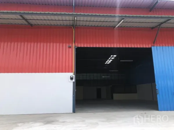 Warehouse for rent in Khlong Luang, Pathum Thani - Close-up of a warehouse bay with a half-open blue sliding door, red metal facade and spacious loading area in front.