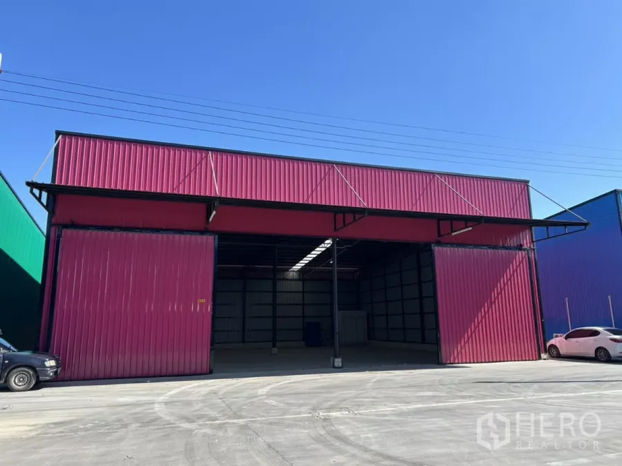 Warehouse for rent in Khlong Toei, Bangkok - Front exterior of a red metal warehouse with wide sliding doors, canopy awnings, and a truck-friendly driveway under a clear sky.