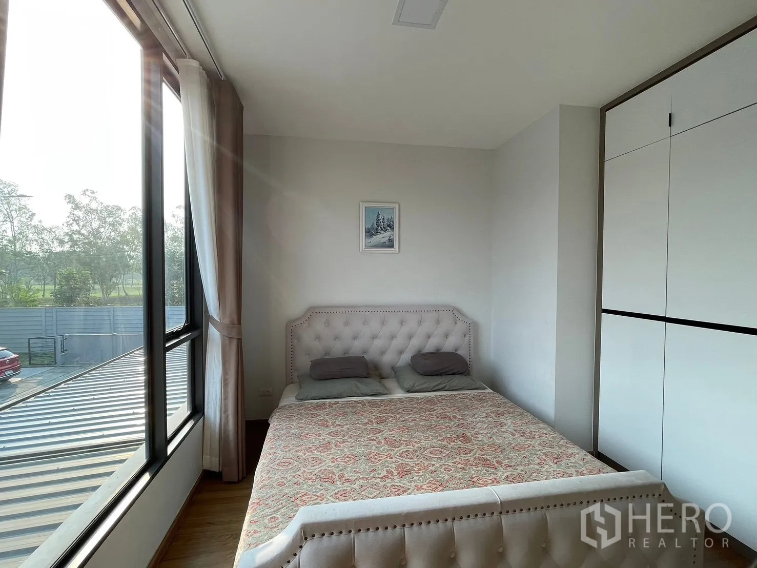 Townhouse for rent in Bang Phli, Samut Prakan - Corner bedroom with large windows overlooking greenery, a tufted bed, and a wardrobe.