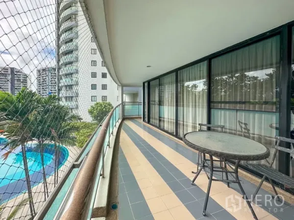 Condominium for sale in Samut Prakan - Wide curved balcony with table overlooking the condo’s swimming pool, palms and neighboring towers.
