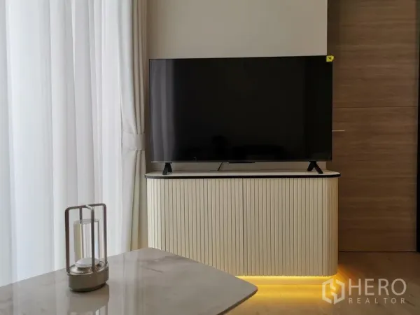 公寓 出租 - 大屏电视置于弧形竖纹柜上，柜底氛围灯柔和，旁为轻薄窗帘。