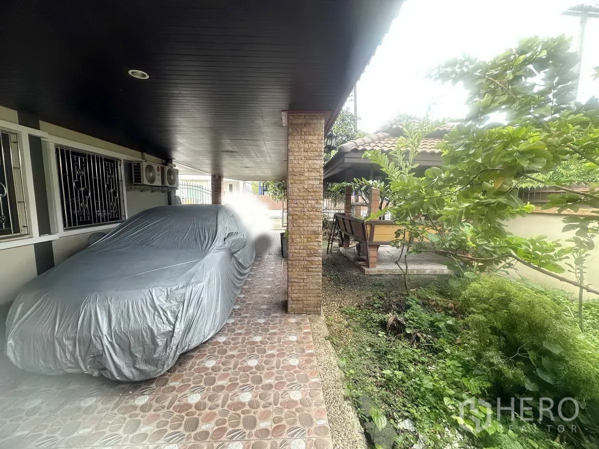 独立式住宅 出售在Pathum Thani - 房侧双车位有顶车棚，旁有绿化与通往凉亭的小径。