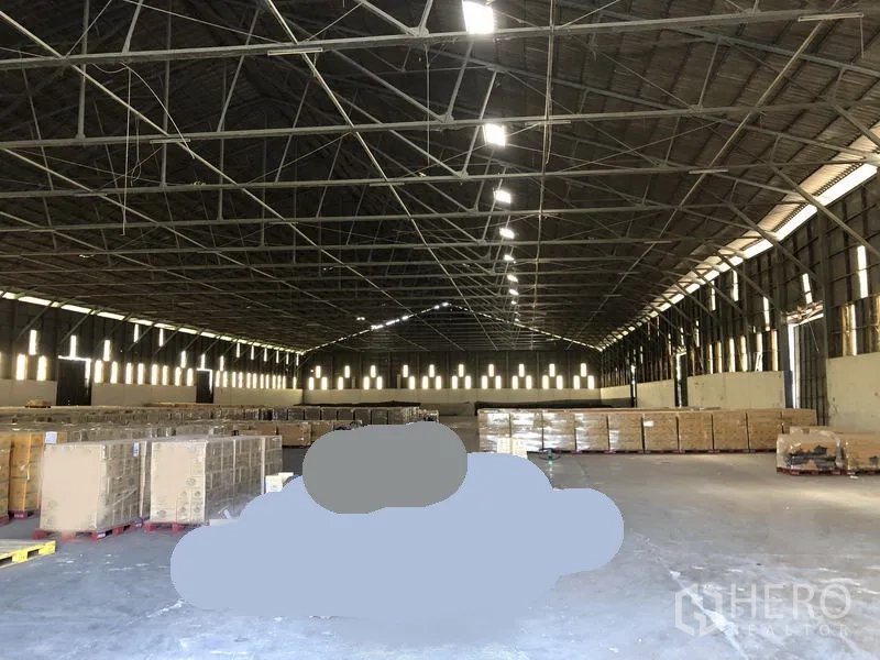 Warehouse for rent in Nonthaburi - Spacious warehouse interior with steel truss roof, skylights and palletized goods.
