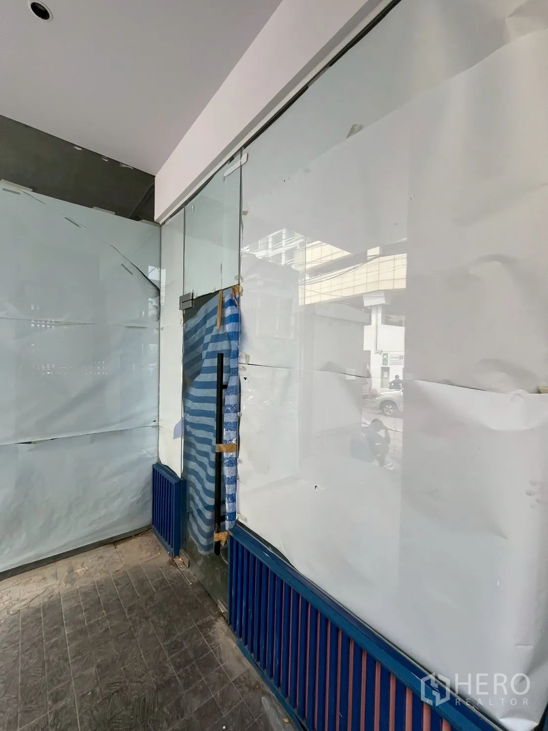 Retail Space for rent in Watthana, Bangkok - Close-up of the glass entrance door to a retail space with temporary coverings, blue and orange slatted base, and tiled flooring reflecting the street outside.