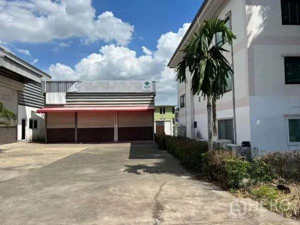 货栈 出租在Prawet, Bangkok - 仓库正面，宽阔混凝土场地、红色雨棚与多扇卷闸门，旁有办公楼与棕榈树，蓝天白云下的景致