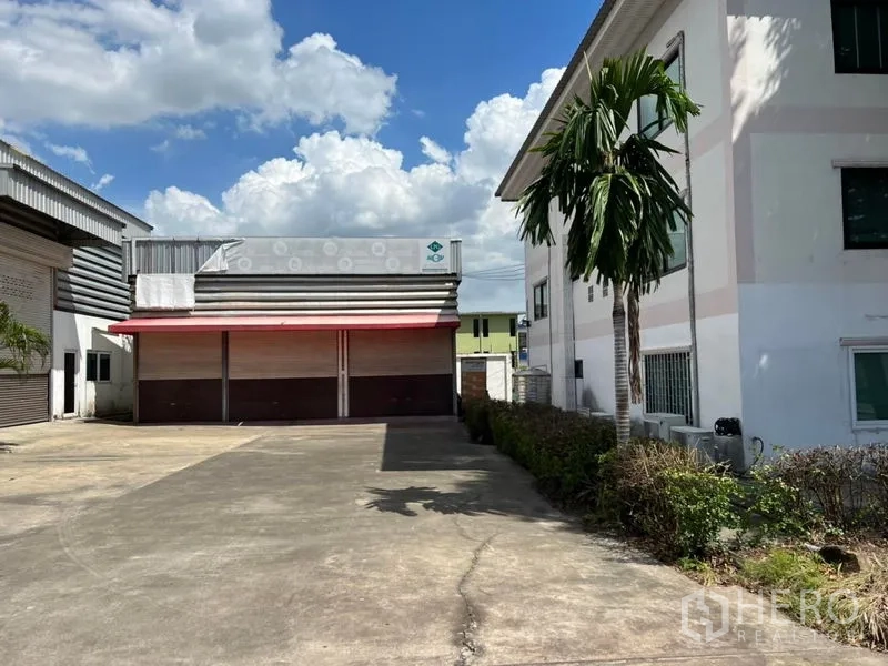 货栈 出租在Prawet, Bangkok - 仓库正面，宽阔混凝土场地、红色雨棚与多扇卷闸门，旁有办公楼与棕榈树，蓝天白云下的景致