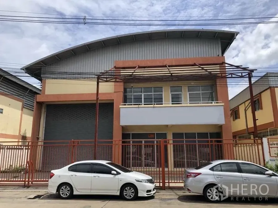 Factory for rent in Pathum Thani - Two-storey factory exterior with curved metal roof, large roller shutter door, front office windows and a secure gated fence with street parking.
