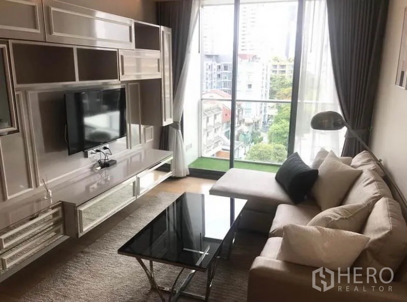 公寓 出租在Watthana, Bangkok - 明亮客厅，带内置电视墙、转角沙发与玻璃茶几，落地窗连接观景阳台。