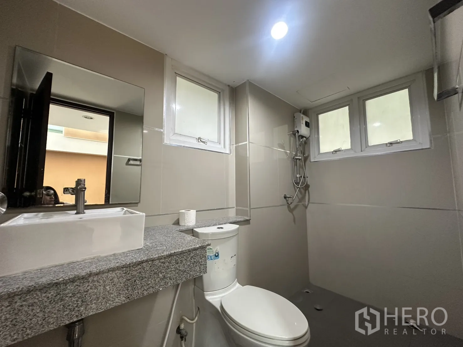 Home Office for rent in Watthana, Bangkok - Compact bathroom with gray tiles, vanity sink, mirror, toilet and a shower with water heater beneath small windows.