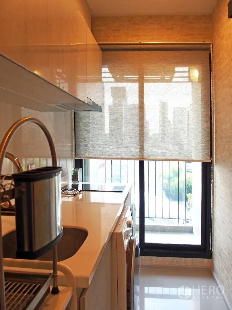 Condominium for rent in Huai Khwang, Bangkok - Compact galley kitchen with sink, induction hob, washing machine, and balcony door with roller blind and city outlook.