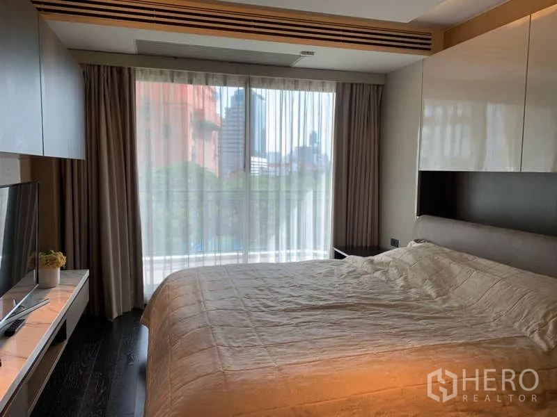 Condominium for rent in Khlong Toei, Bangkok - Cozy bedroom with large bed, TV console and city view through sheer curtains.