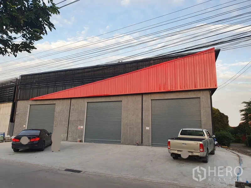 Warehouse for rent in Pak Kret, Nonthaburi - Front elevation showing three large grey roller shutter doors with parking on a broad concrete forecourt.