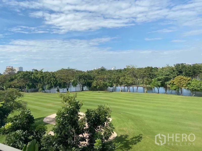 Townhouse for rent or sale in Bang Kapi, Bangkok - Manicured green lawn and lakeside view with trees and blue sky near the community.