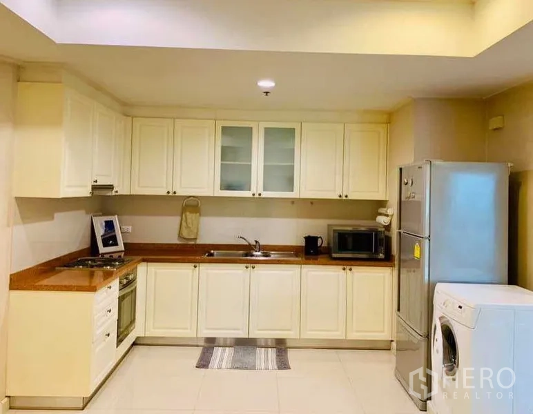 Condominium for rent in Khlong Toei, Bangkok - Spacious L-shaped kitchen with white cabinets, built-in hob and oven, fridge and washing machine.