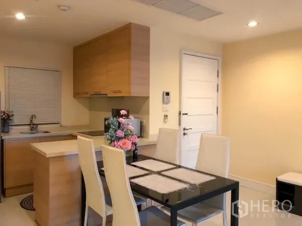 คอนโด ให้เช่า - ครัวบิลท์อินไม้พร้อมอ่างล้างจานและตู้ลอย ข้างโต๊ะทานอาหาร 4 ที่นั่ง ใกล้ประตูสีขาว