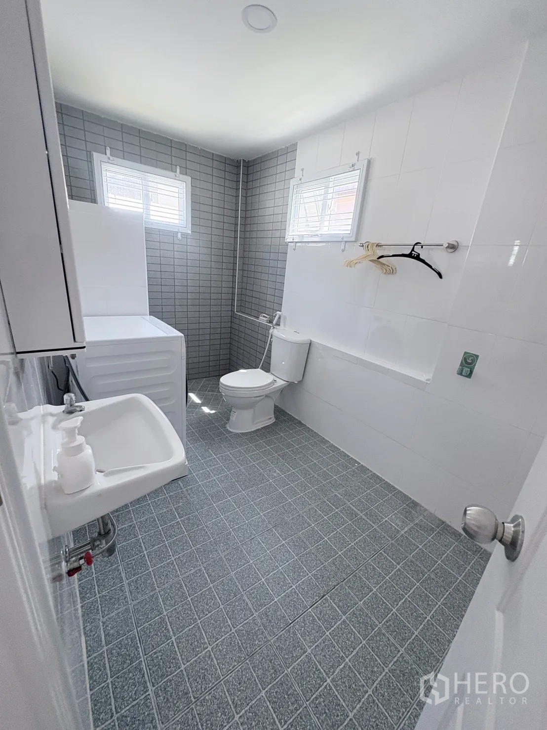 Detached House for rent in Saphan Sung, Bangkok - Bright bathroom with gray tiles, two windows, toilet, sink, washing machine, and shower.