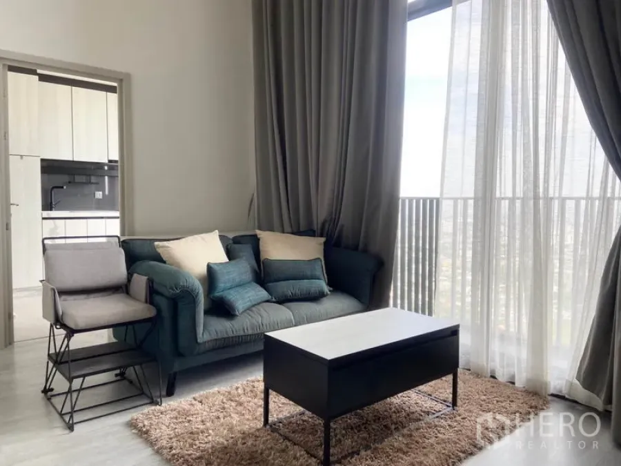 Condominium for rent in Phra Khanong, Bangkok - Bright living room with teal sofa, tall curtains, floor-to-ceiling windows and a doorway to the kitchen with city views beyond.