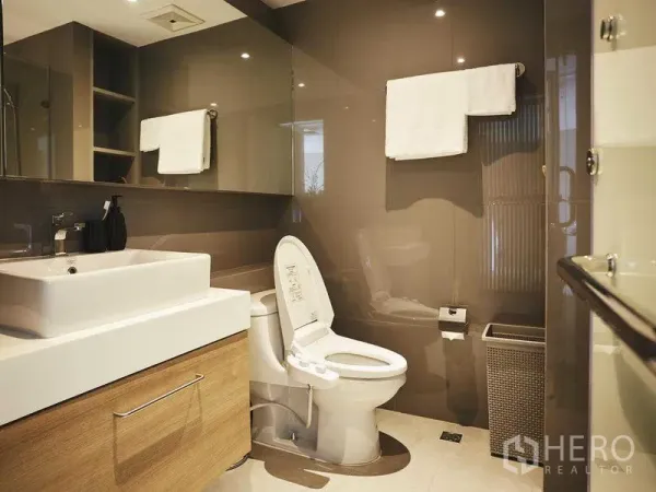 Apartment for rent in Khlong Toei, Bangkok - Modern bathroom with glossy wall panels, square basin vanity, mirror cabinets and toilet in neutral tones.