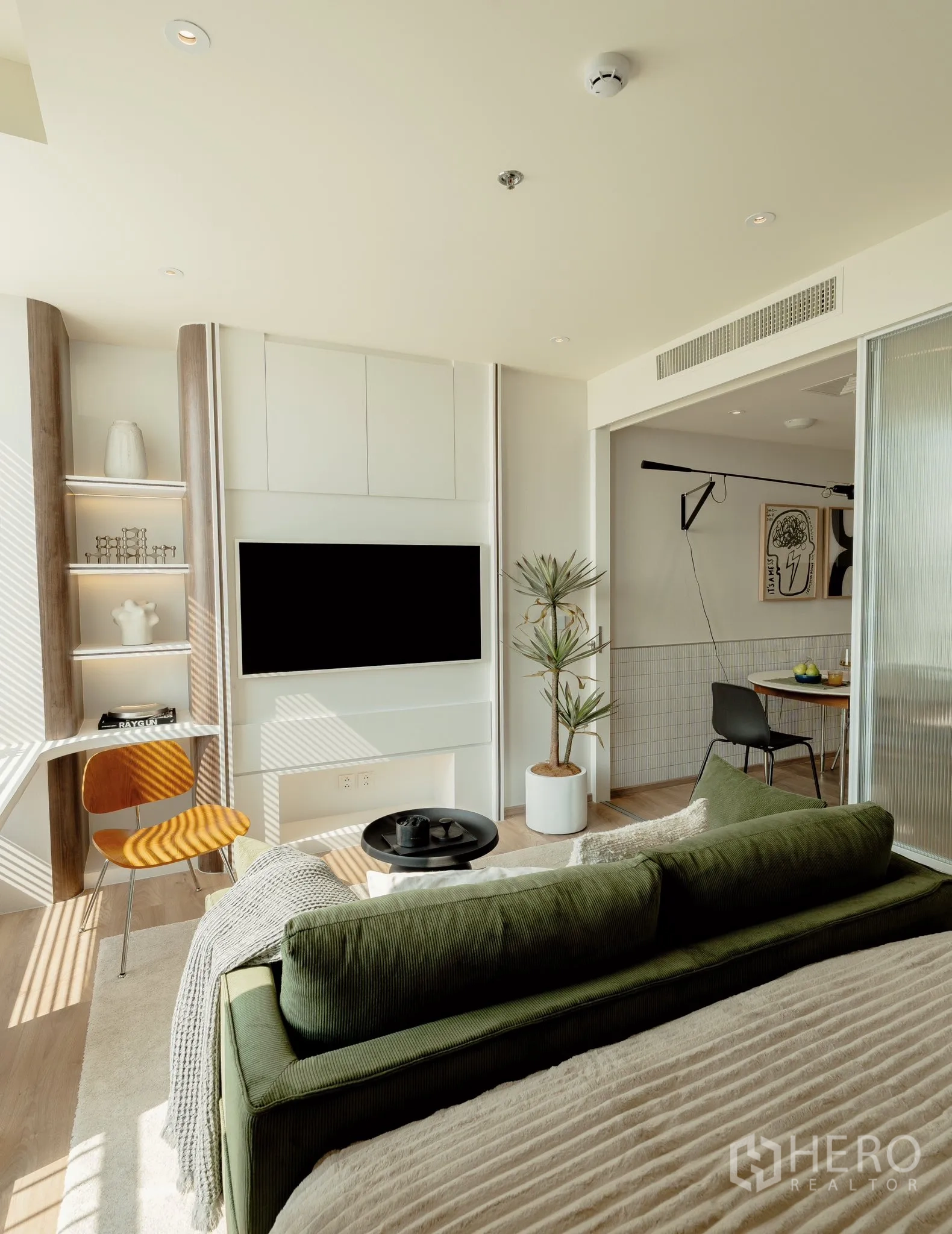 Condominium for rent in Watthana, Bangkok - Sunlit living-sleeping area with green sofa and bed, built-in TV wall with shelves, small desk, potted plant, and a sliding glass door to the dining kitchen.